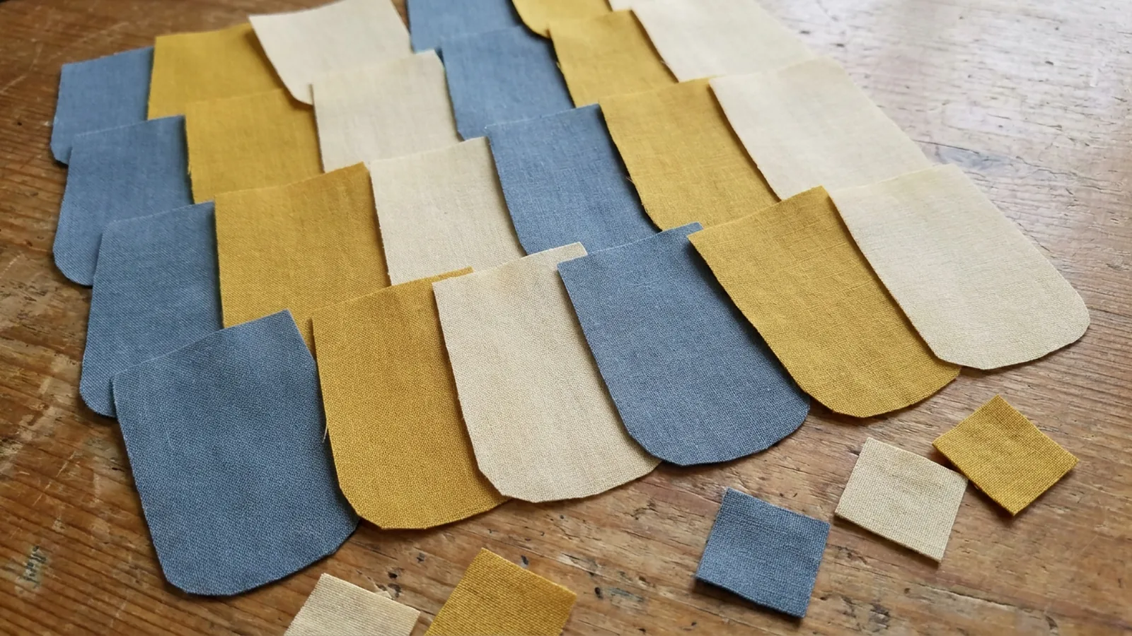 Illustrative generated image of tumbler-shaped cotton pieces in blue, mustard, and cream on a wooden table, with a few small squares nearby. Not a photograph of a finished quilt.